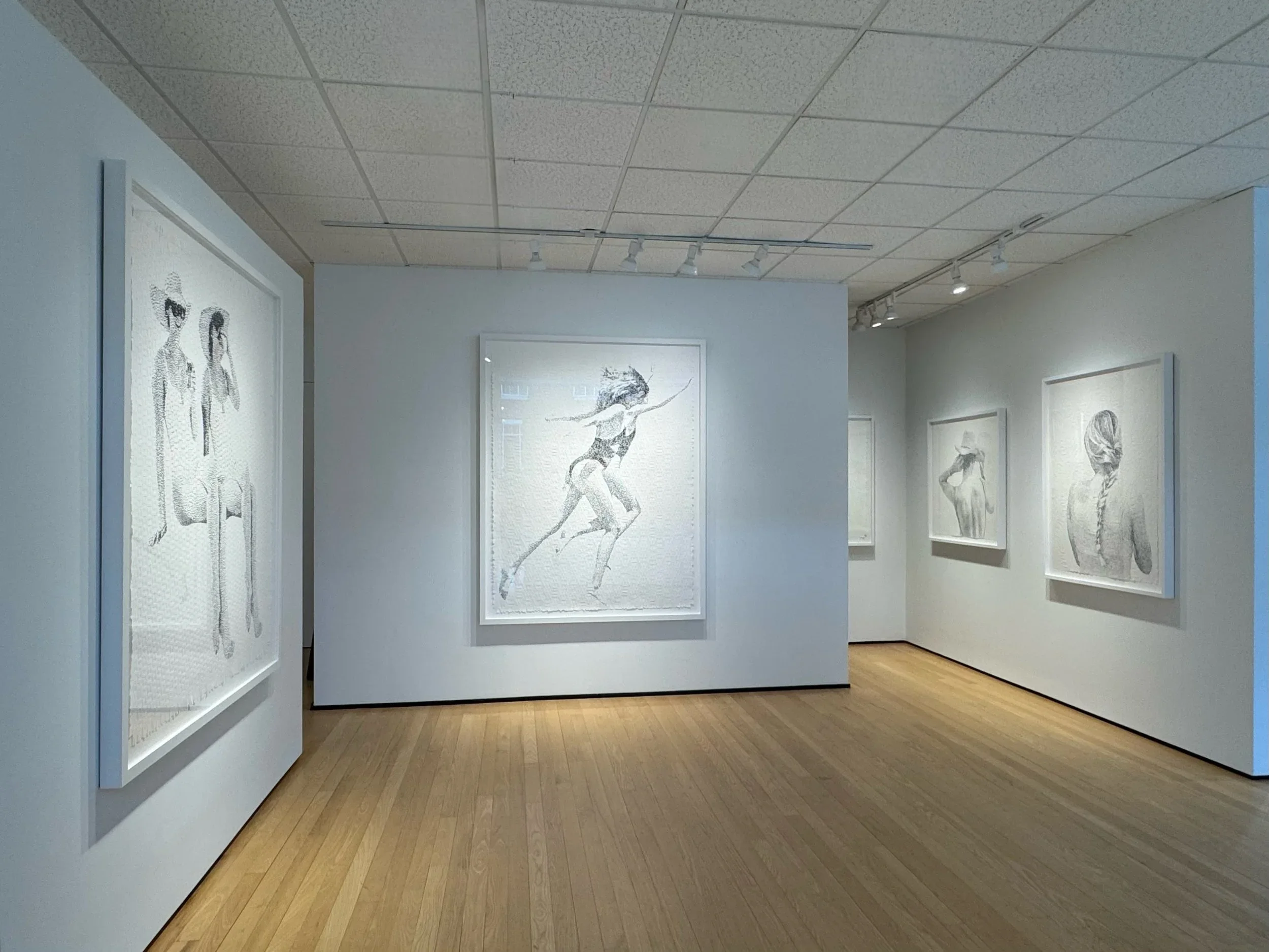 Art gallery with five black and white sketches of women on white walls, wooden floor, and ceiling with track lighting.