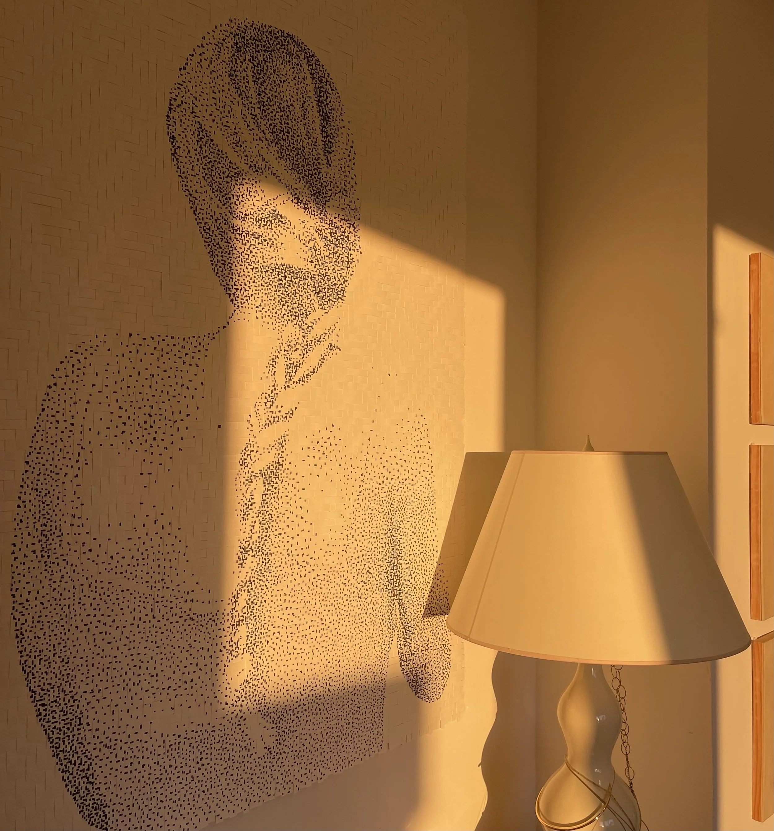 A pixelated portrait of a woman with short hair, cast in warm sunlight, standing next to a lamp with a cream-colored shade in a room.
