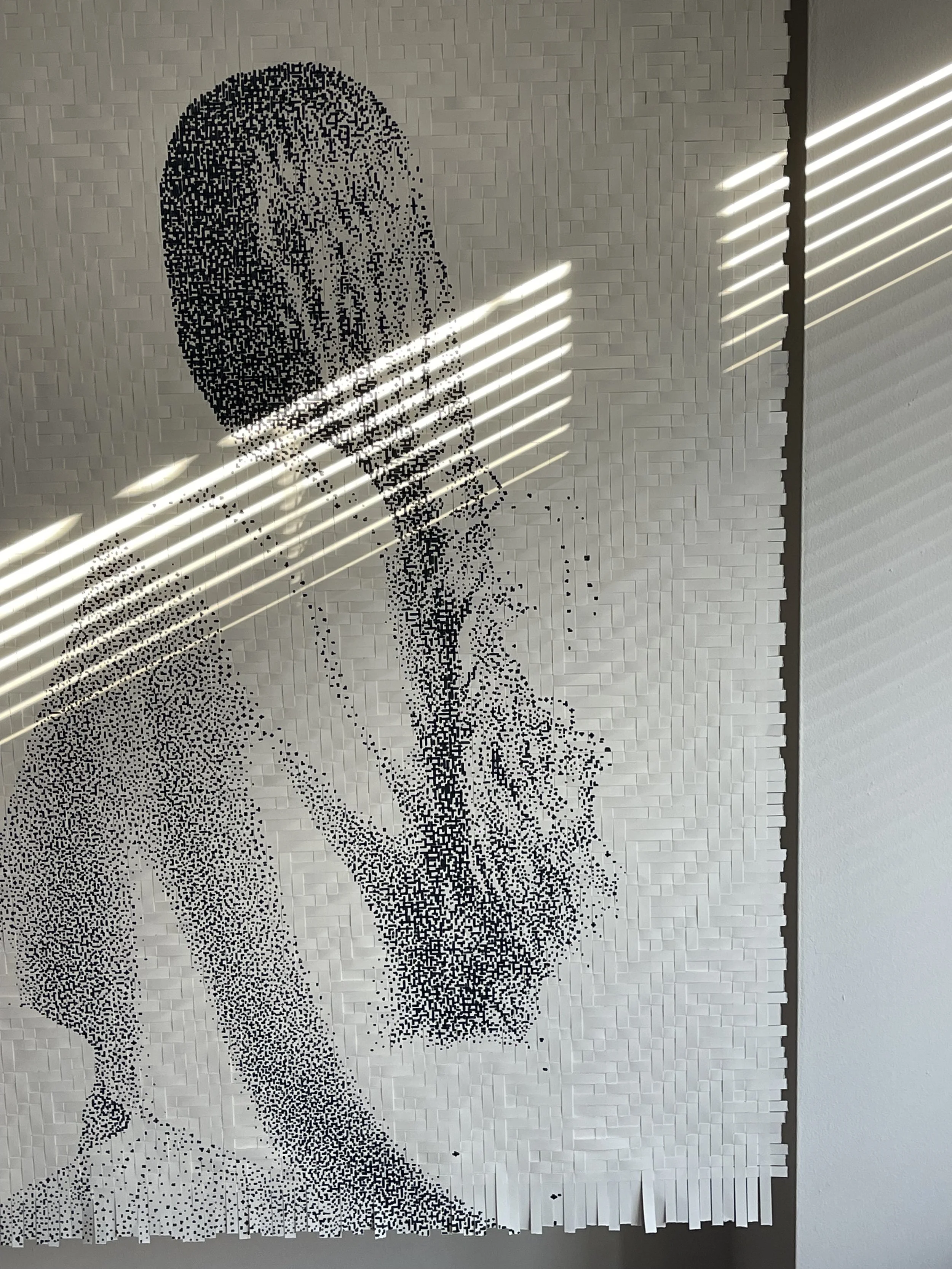 Black and white pixel art of a woman resting her chin on her hand, with her hair in a bun, created from small paper pieces on a wall.