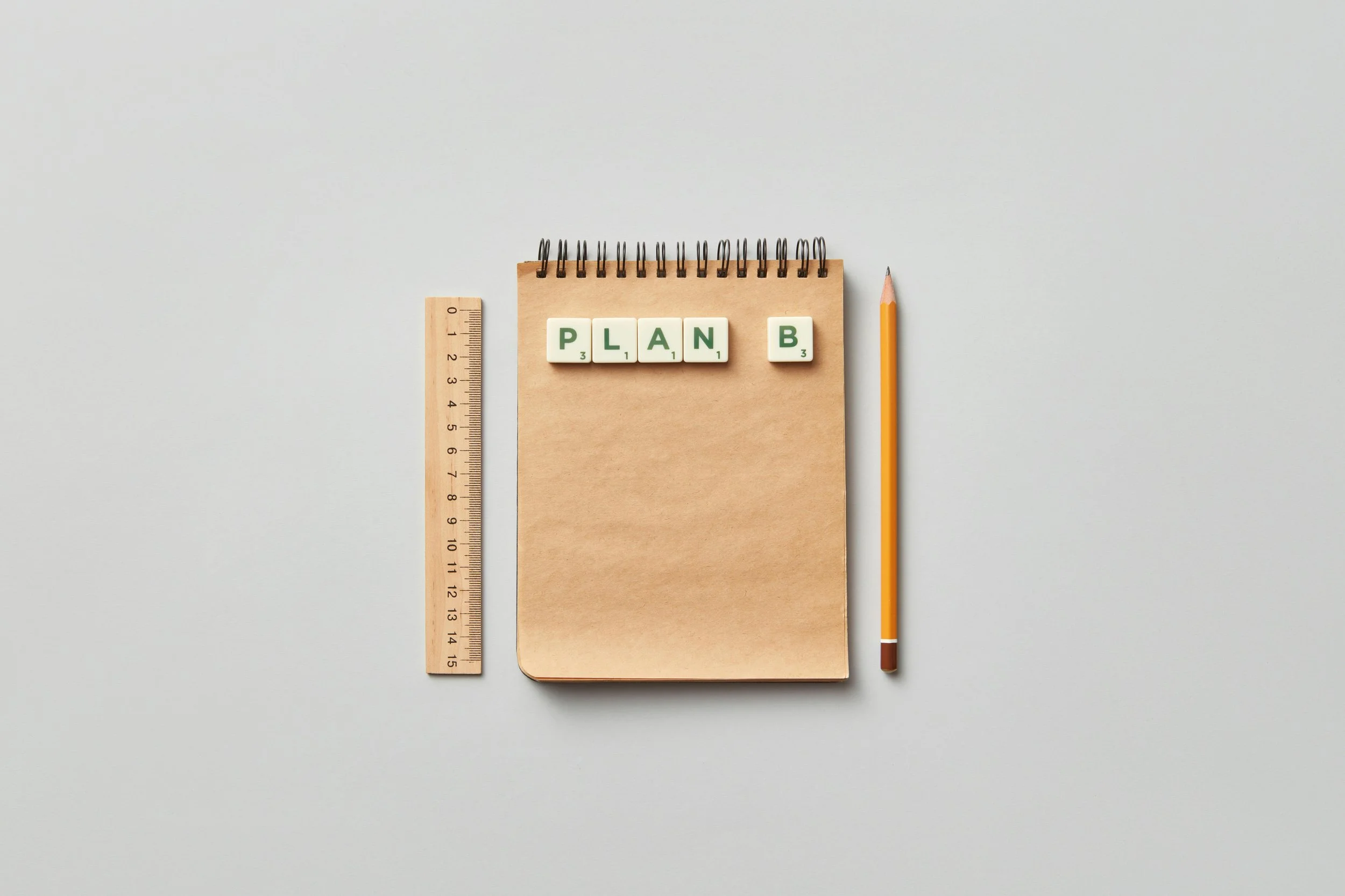 Notebook with Scrabble tiles spelling 'PLAN B', a ruler, and a pencil on a light gray surface.