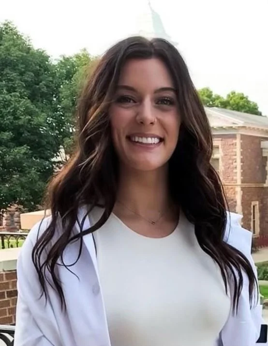 Young woman with long brown hair smiling outdoors, wearing a white top and jacket, with trees and a brick building in the background.