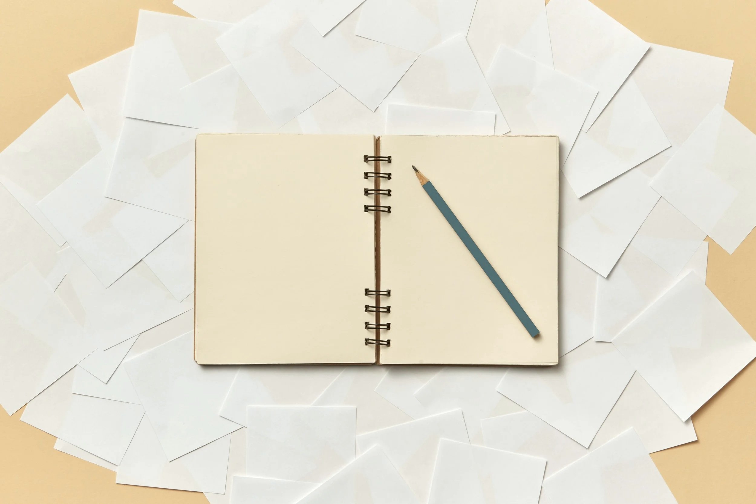 An open blank notebook with a blue pencil resting on it, surrounded by scattered white envelopes on a light beige surface.
