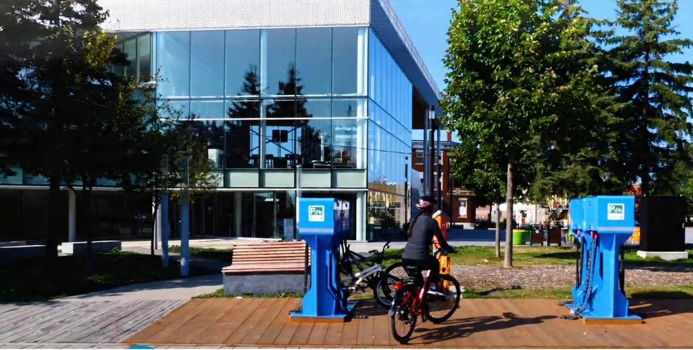A person riding a bicycle past blue bicycle parking stations in an urban park with trees and a modern glass building.