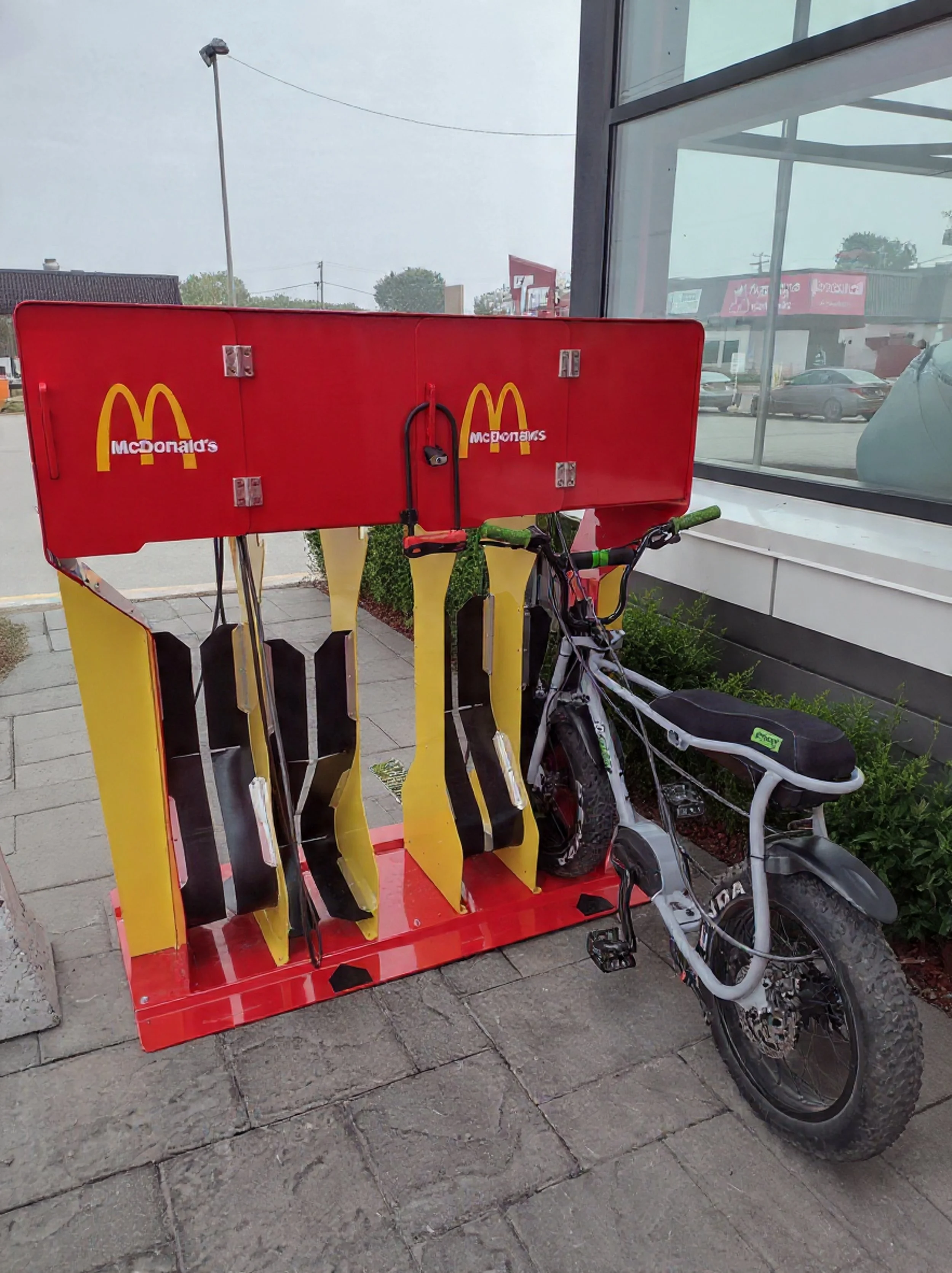 A yellow and red McDonald's-branded bike rack with a black bicycle attached outside a restaurant, with windows reflecting a street scene.