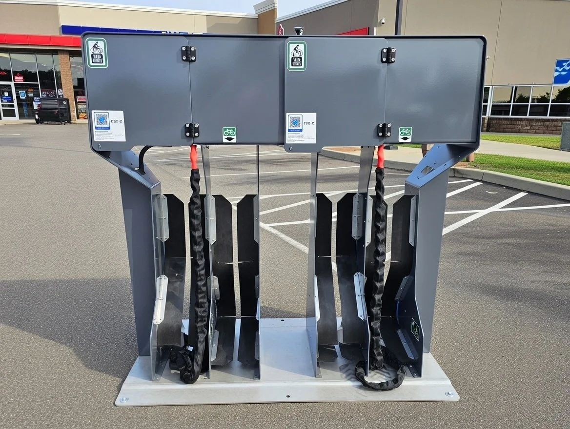 Outdoor bike rental station with three docking spots, each equipped with a security strap, located in a parking lot outside a commercial building.