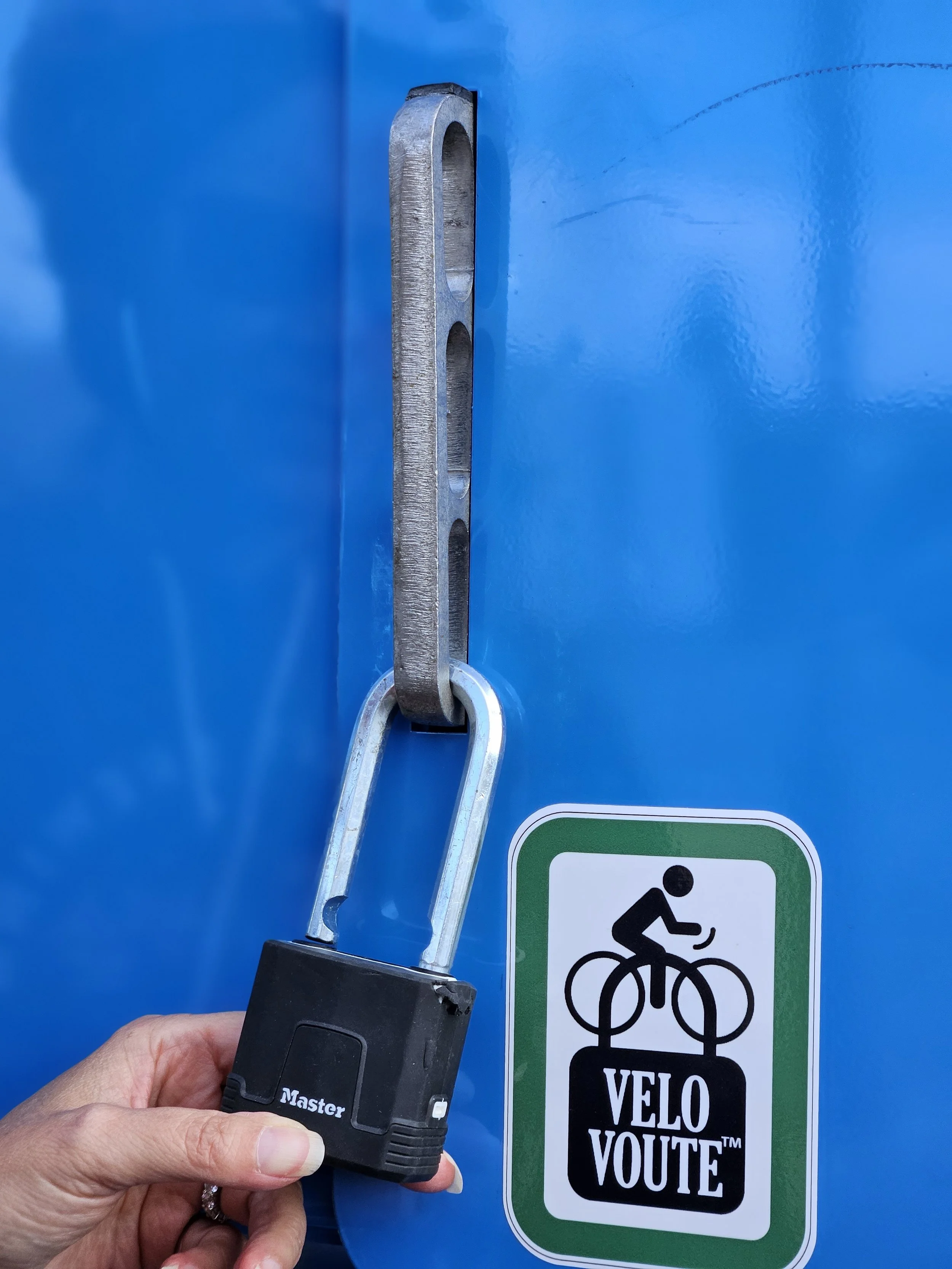 A close-up of a blue bike lock attached to a sign that says 'VELO VOUTE' with a silhouette of a cyclist. The lock is being held by a person's hand.