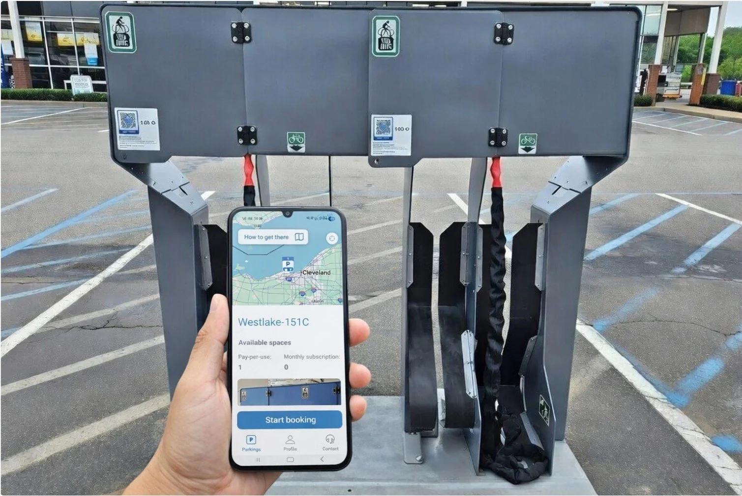 A person holds a smartphone displaying a parking app with available spaces in a parking lot. Behind, there is a motorized bike rack with two sections, each with a bike lock, situated in an outdoor parking area with painted parking lines and a buildin