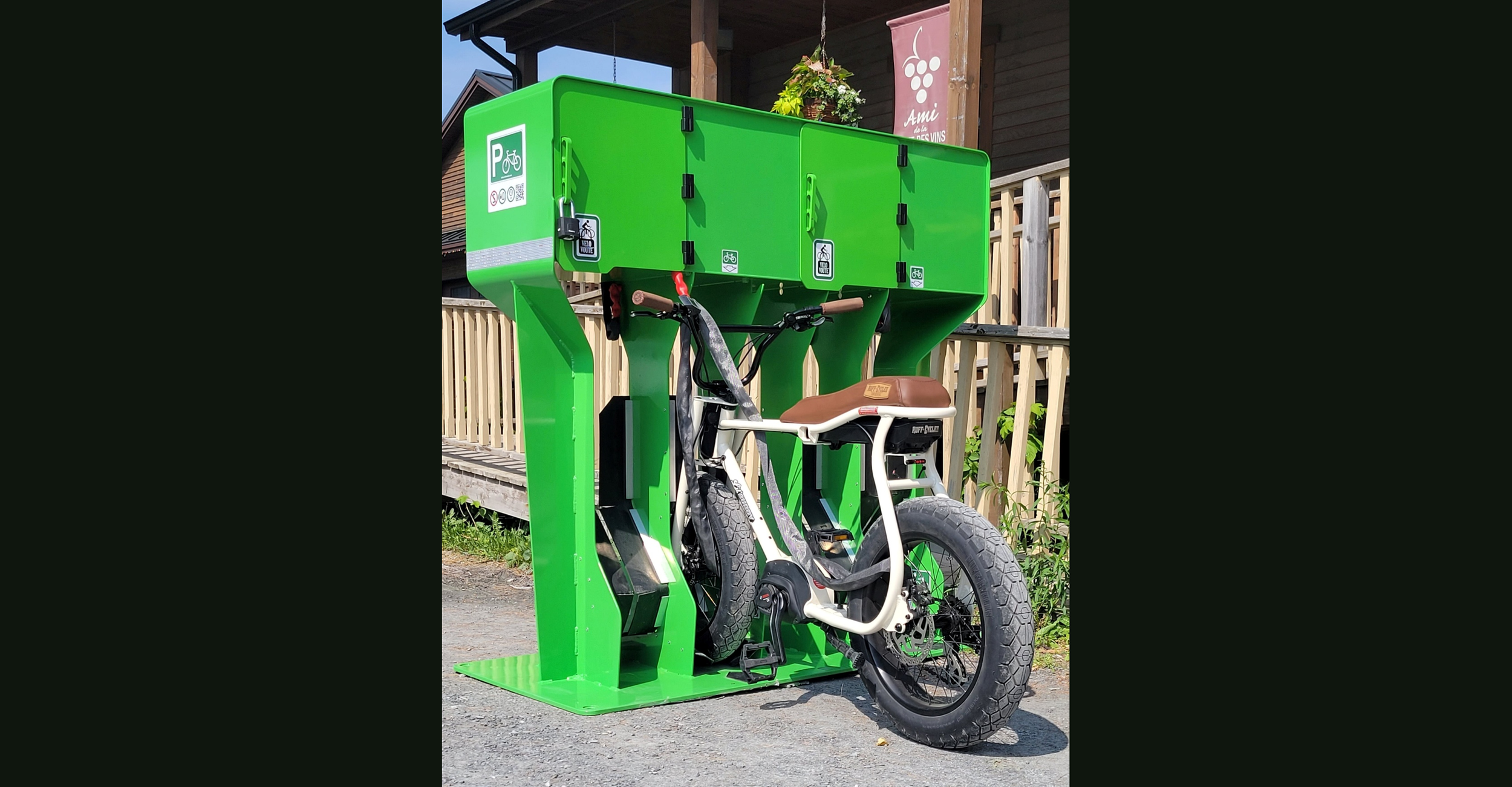 Green bike-sharing station with an e-bike parked in front of it, located outdoors on a paved surface with wooden fencing and a building with a balcony and hanging flower basket in the background.
