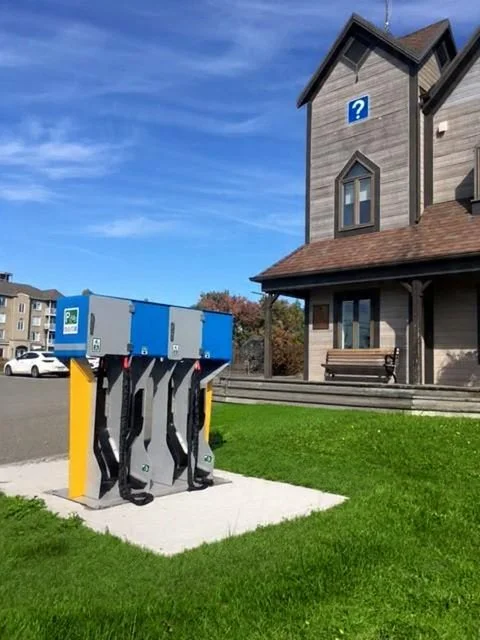 An electric vehicle charging station with three ports located outside a building with a question mark symbol on the door and above the window, with a grassy area and a bench.