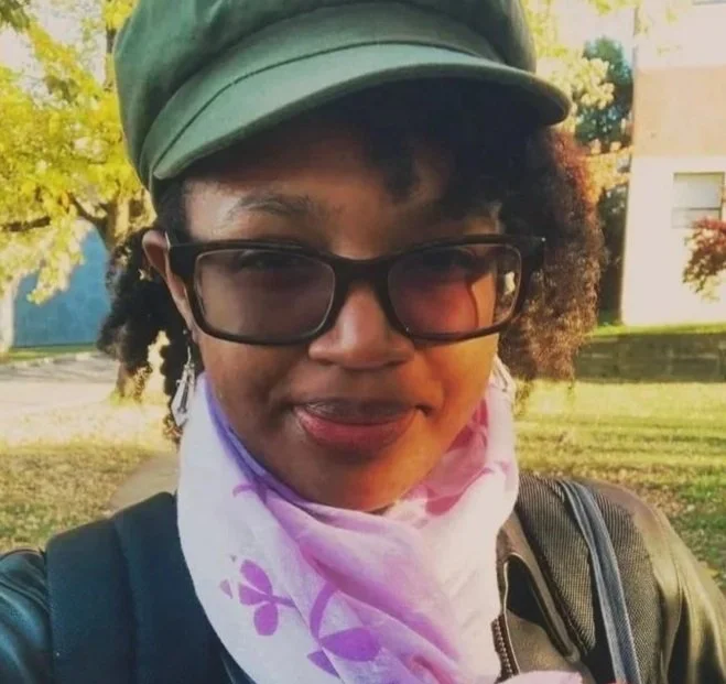 A close-up of a young woman outdoors wearing glasses, a green cap, a black jacket, and a pink scarf with purple flowers, smiling at the camera.