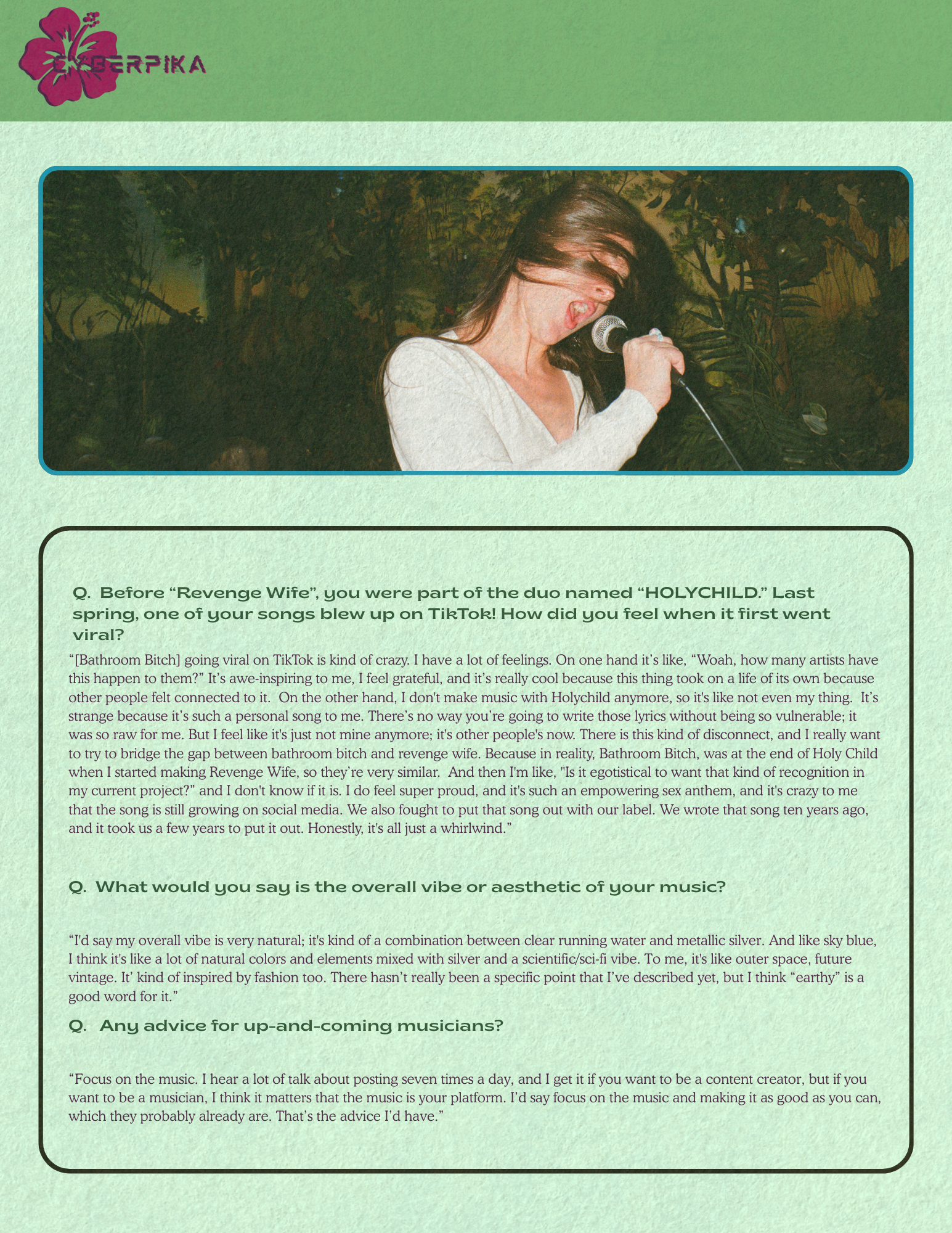 A woman singing into a microphone with long hair blowing in the wind, background appears to be a lush, green indoor setting with tropical plants.