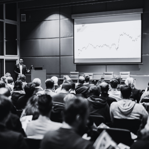 A speaker presents at a conference with a projected line graph showing upward trends, audience members listening attentively.