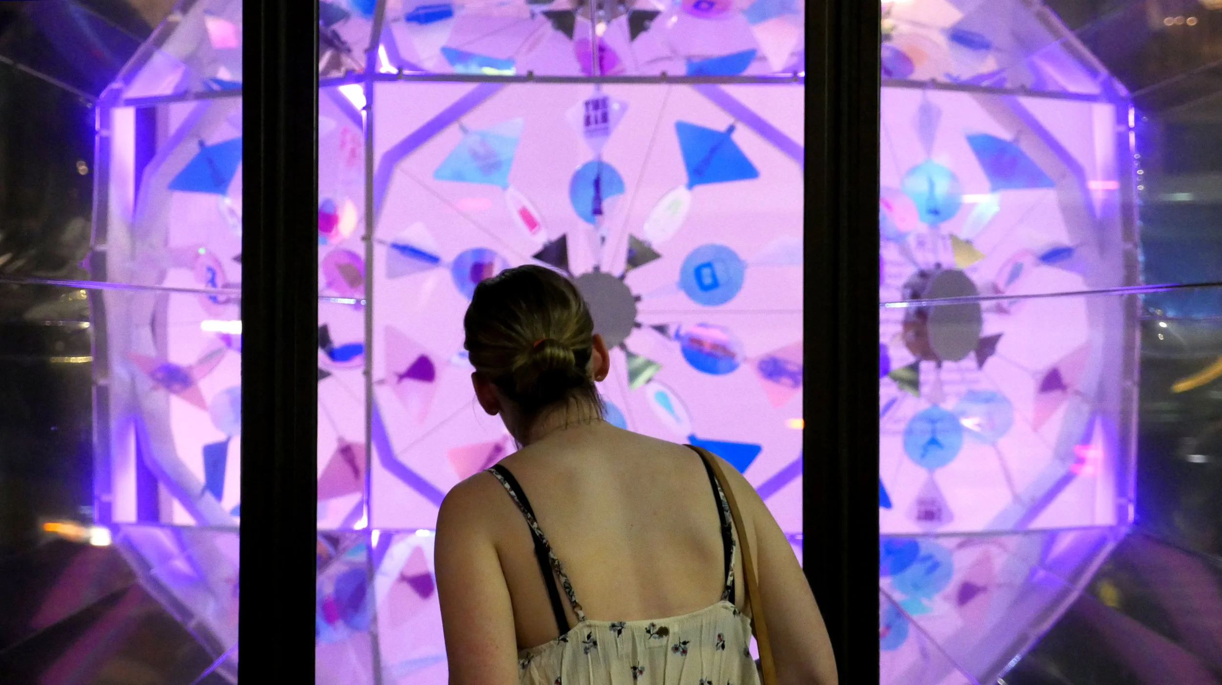 A woman with a ponytail, wearing a sleeveless top, looks at a colorful, illuminated wheel with app icons in a dark environment.