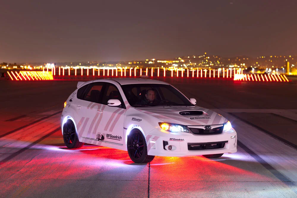 A white Subaru racing or testing vehicle with BFGoodrich branding, illuminated by underglow lights, on a runway or open area at night with city lights in the background.