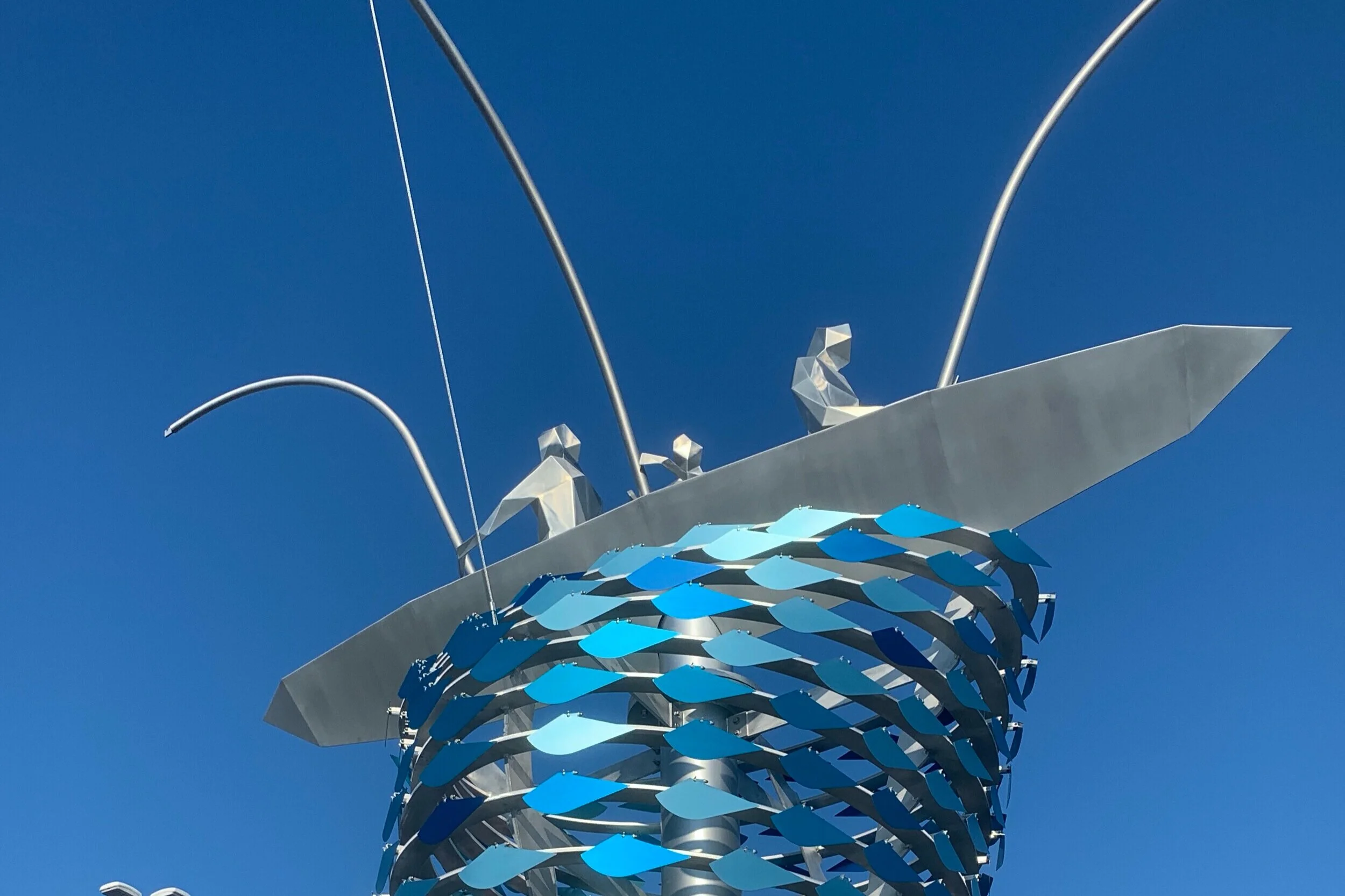 Artistic metal sculpture resembling a fish with water-like blue scales, set against a clear blue sky.