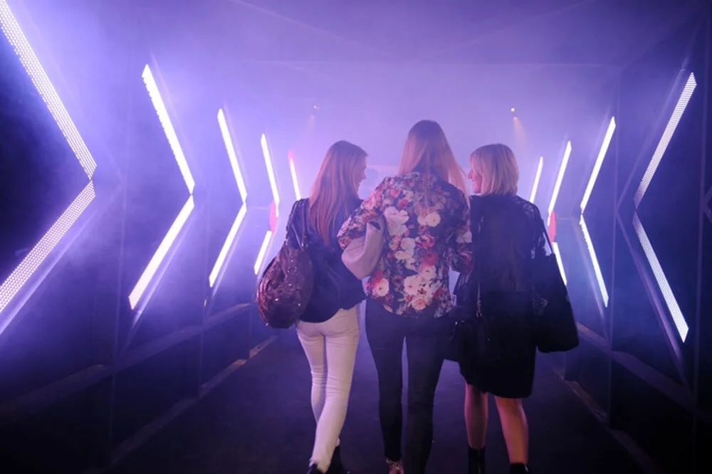 Three women walking through a purple-lit tunnel with futuristic LED light panels.