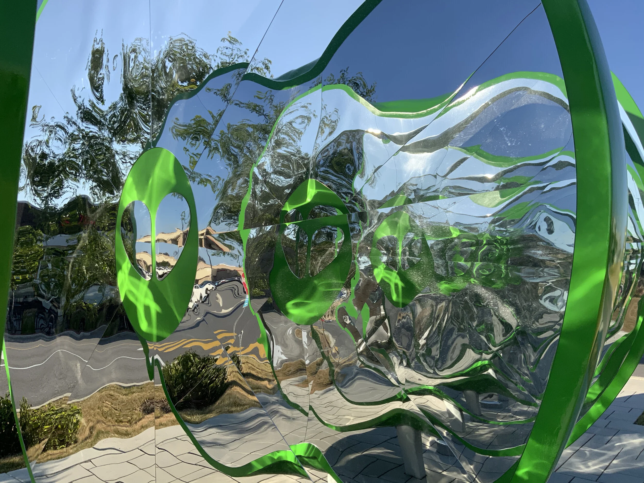 Reflective outdoor sculpture with green and silver distorted shapes, surrounded by trees and a clear blue sky.