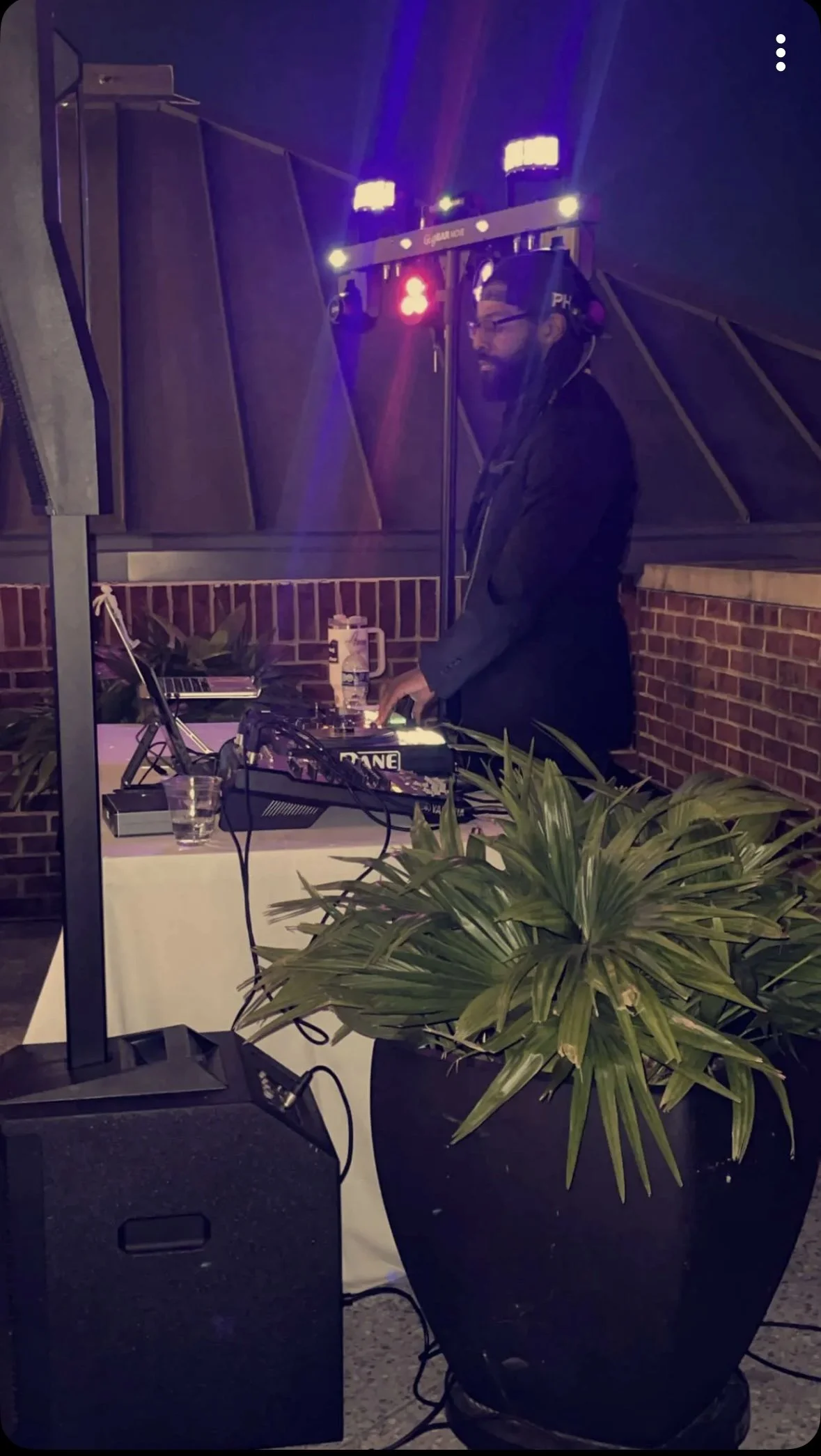 A man DJing at an outdoor event at night, wearing headphones and a blazer, with sound equipment and plants around him.