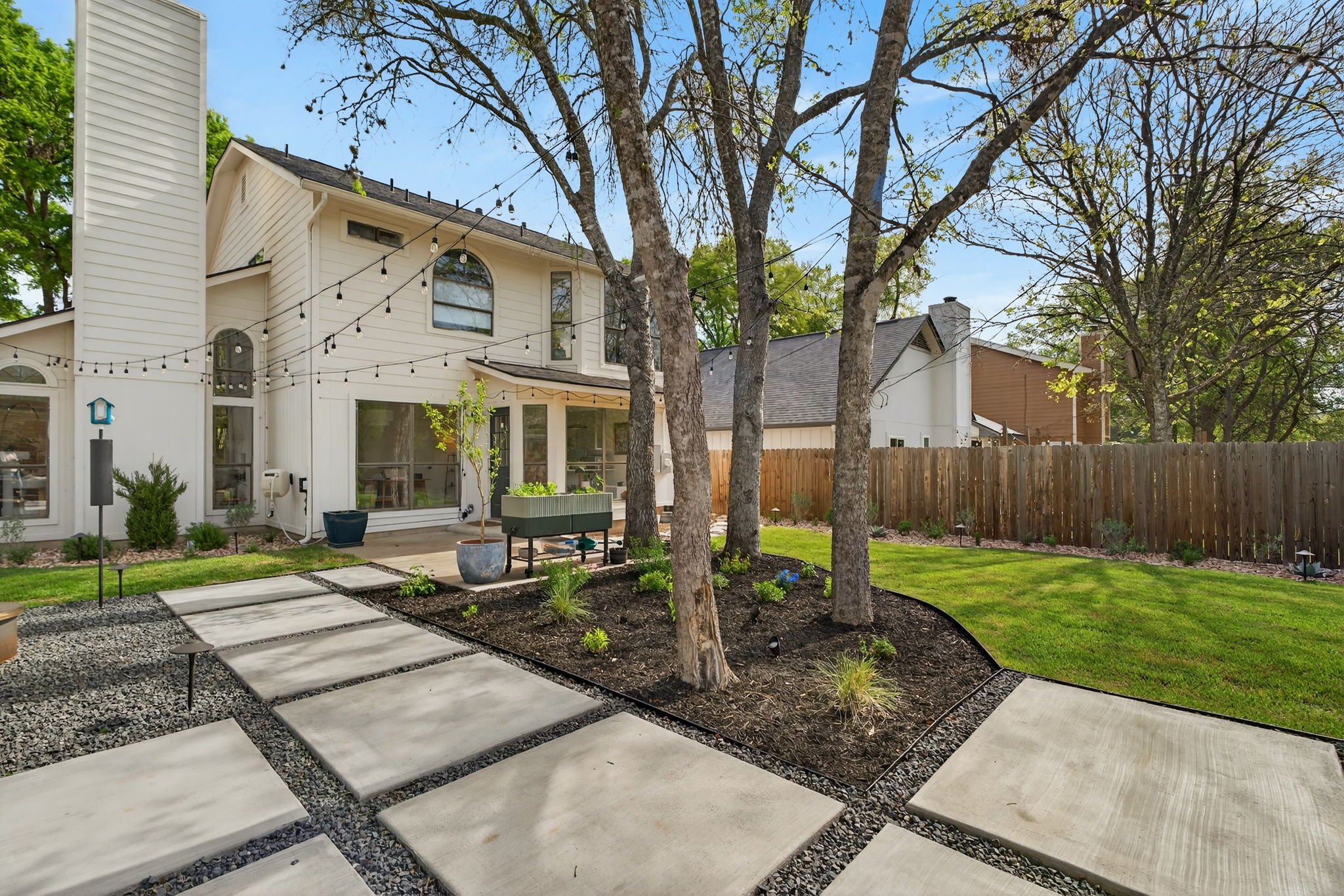 Backyard with concrete paver patio, gravel joints, mulched tree bed, and string lights over the lawn