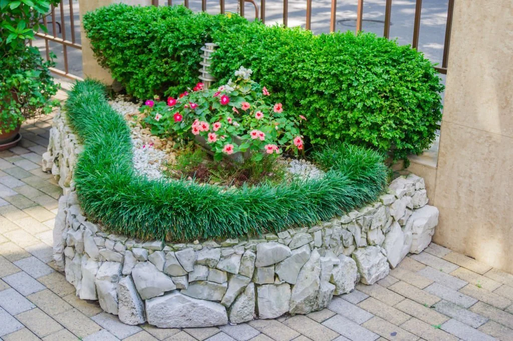 Curved white stone retaining wall surrounds a vibrant flower bed with pink blooms and dense hedges for added landscape support.