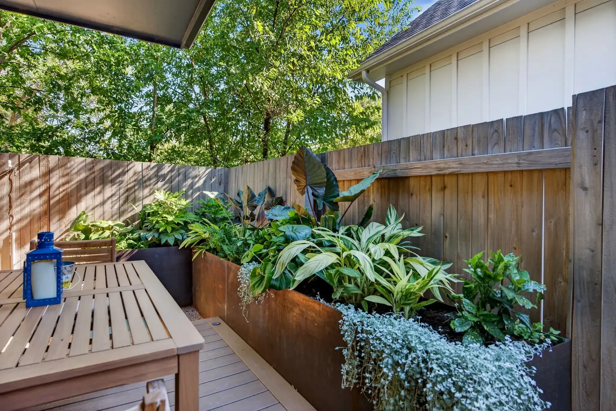 Modern backyard with wooden privacy fence and large planter box filled with tropical foliage beside a cozy dining area.