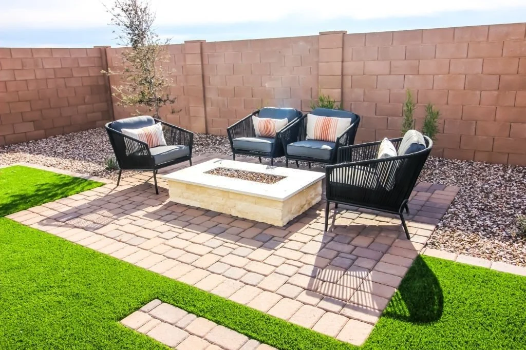 Modern backyard patio with black wicker chairs, a stone fire pit, paver flooring, artificial grass, and a brick privacy wall.