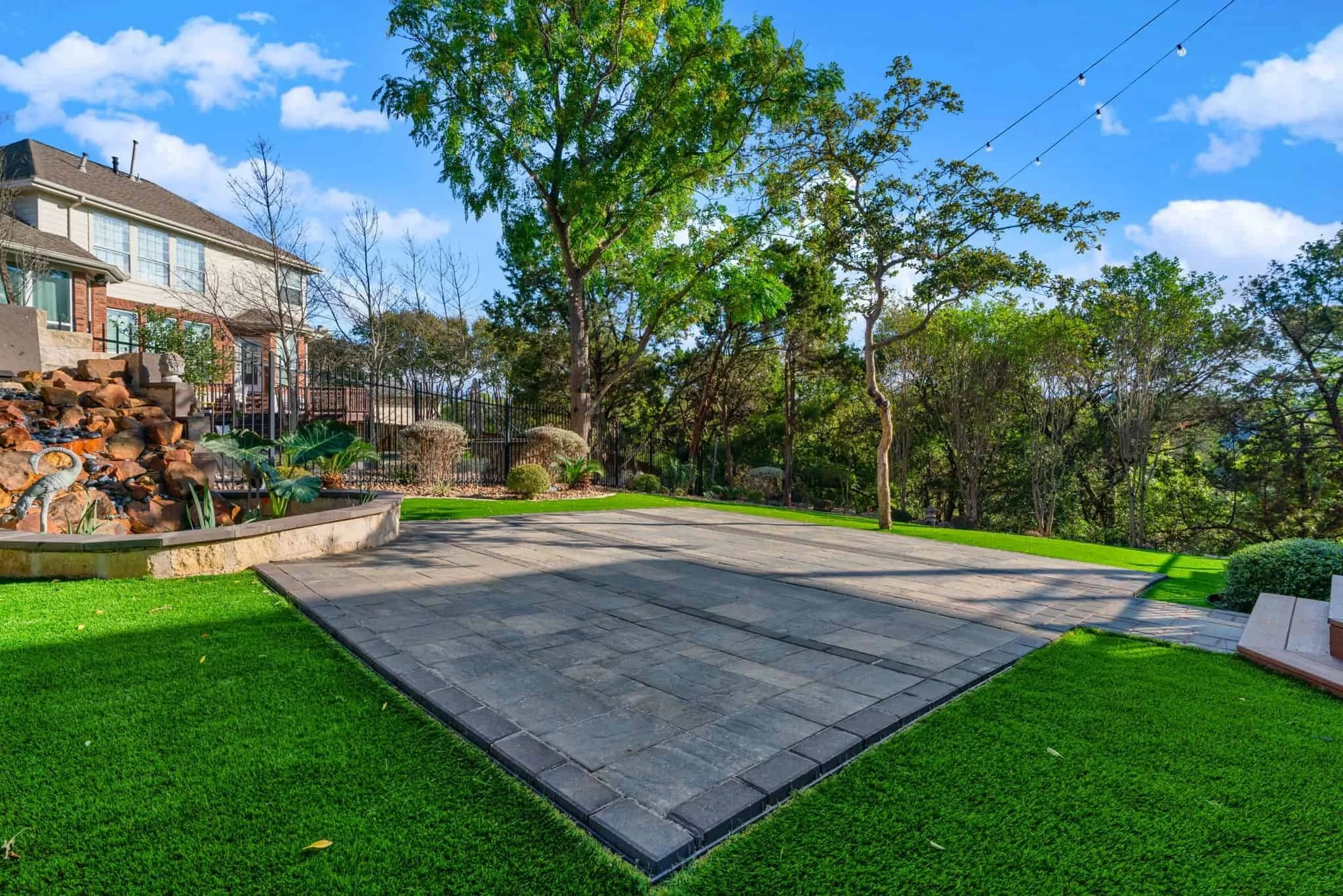 Modern patio with square paver platform, artificial turf, large tree canopy, and a cascading rock water feature under a blue sky.