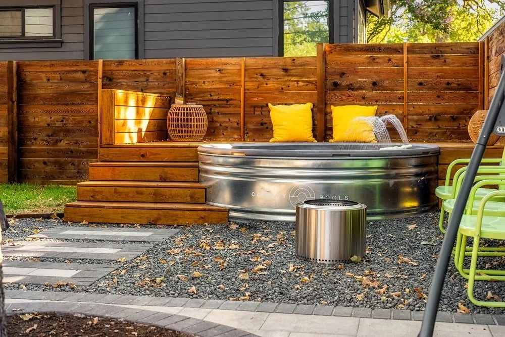Backyard with a stock tank pool, cascading water feature, wood deck, and yellow pillows, enclosed by a wooden fence.
