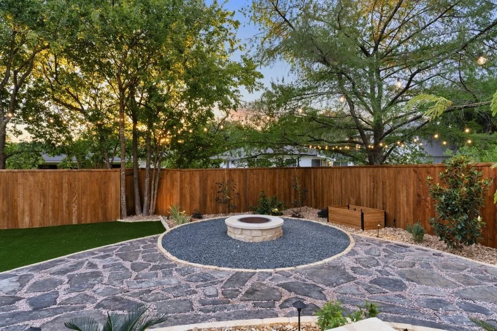 Round stone firepit surrounded by gravel and flagstone patio, string lights, and wooden fence in landscaped backyard with trees.