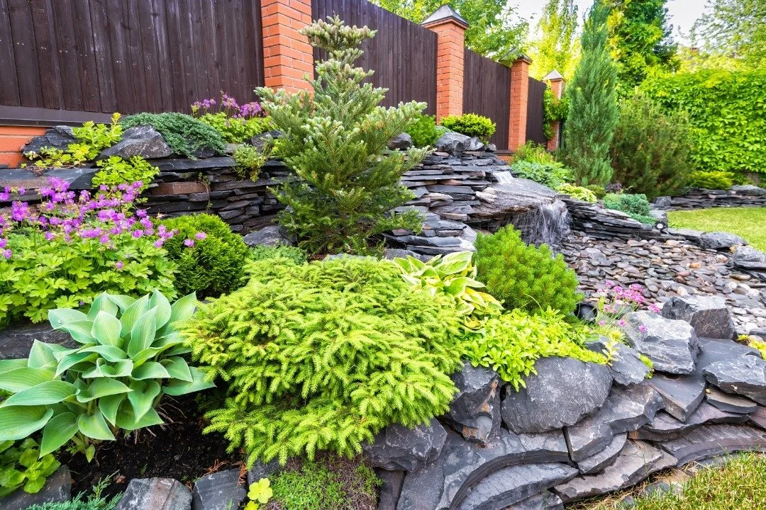Tiered rock garden with a cascading waterfall, colorful flowers, and lush greenery, bordered by a dark wood and brick fence.
