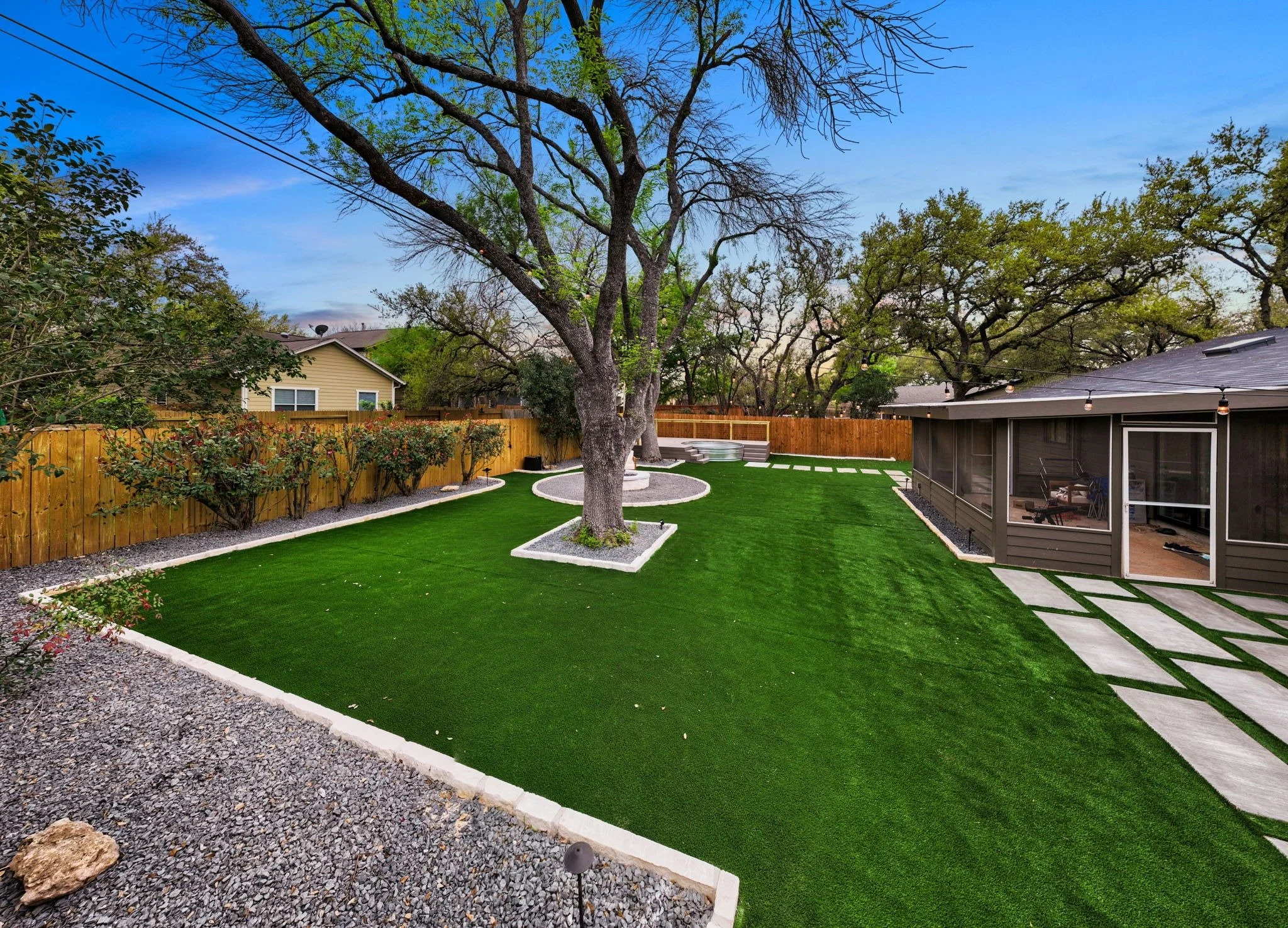 Spacious backyard with artificial grass, a fire pit area, a screened patio, and mature trees, enclosed by a wooden fence.