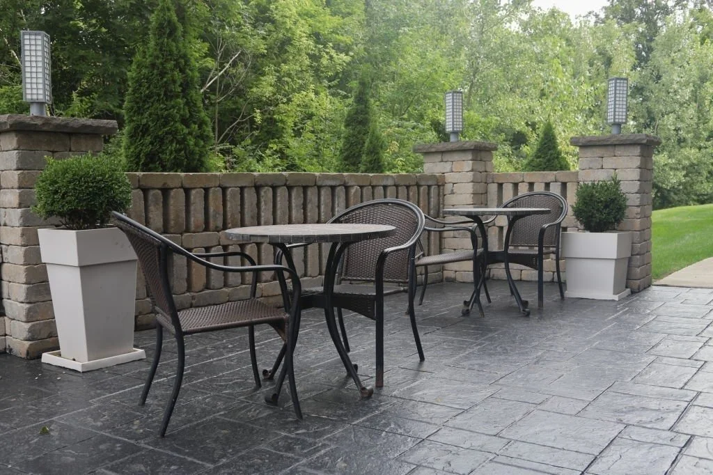 Elegant stamped concrete patio with wrought iron furniture, stone pillars, planters, and lush greenery in a secluded backyard.