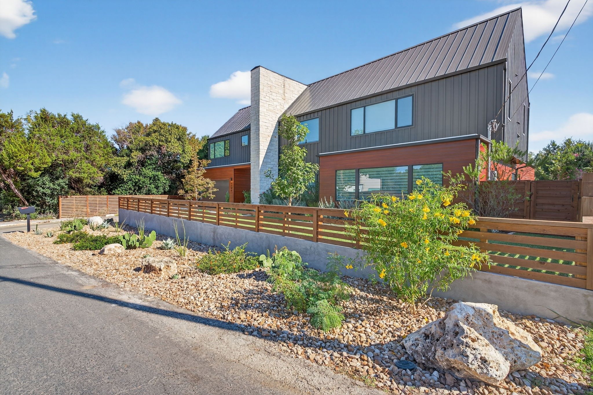 Modern home with desert-style landscaping, wood fence, drought-tolerant plants, and gravel-filled front yard with large rocks.