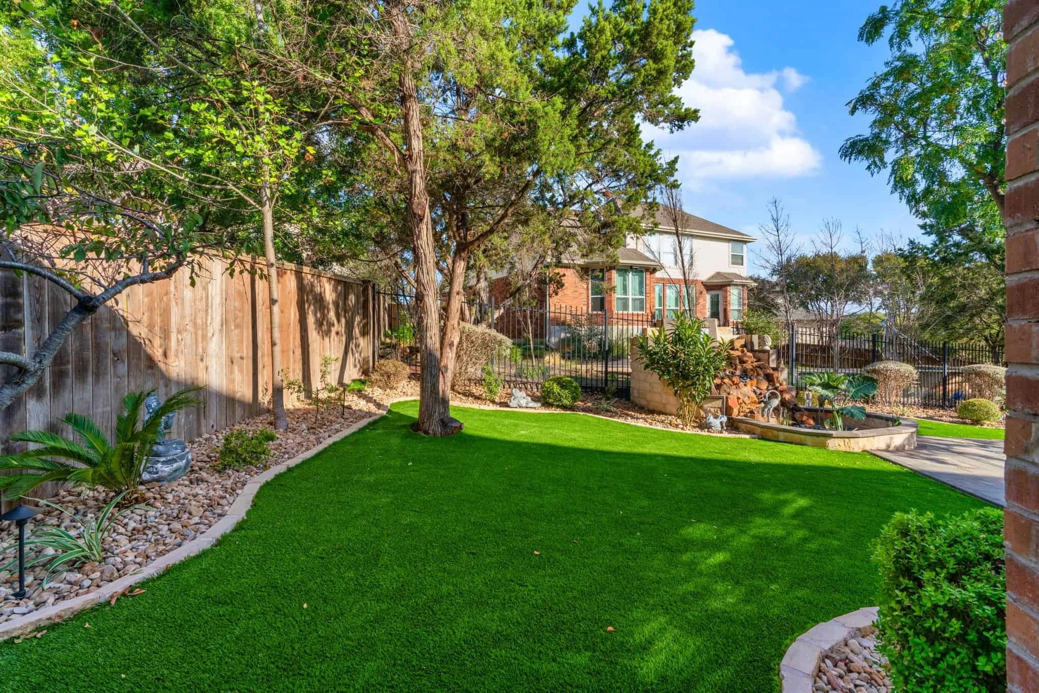 Landscaped backyard with synthetic grass, curved stone edging, mature trees, decorative rocks, and a tiered water feature.