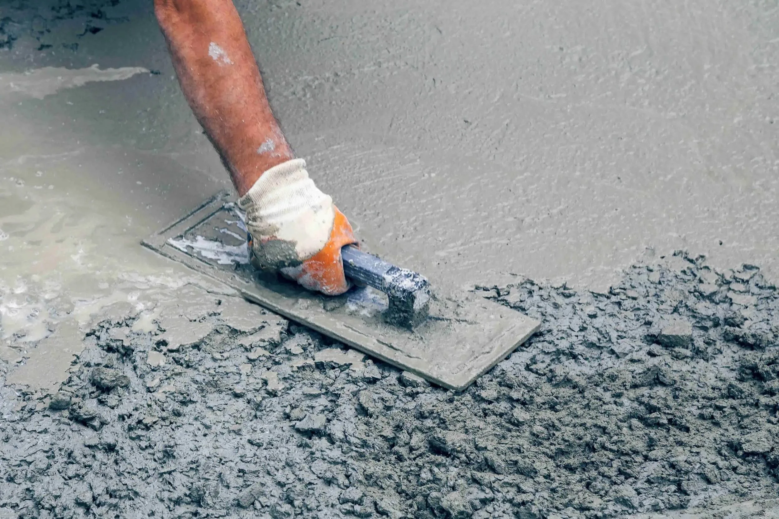 A construction worker in gloves smooths wet concrete with a trowel, leveling the rough surface during a masonry project.