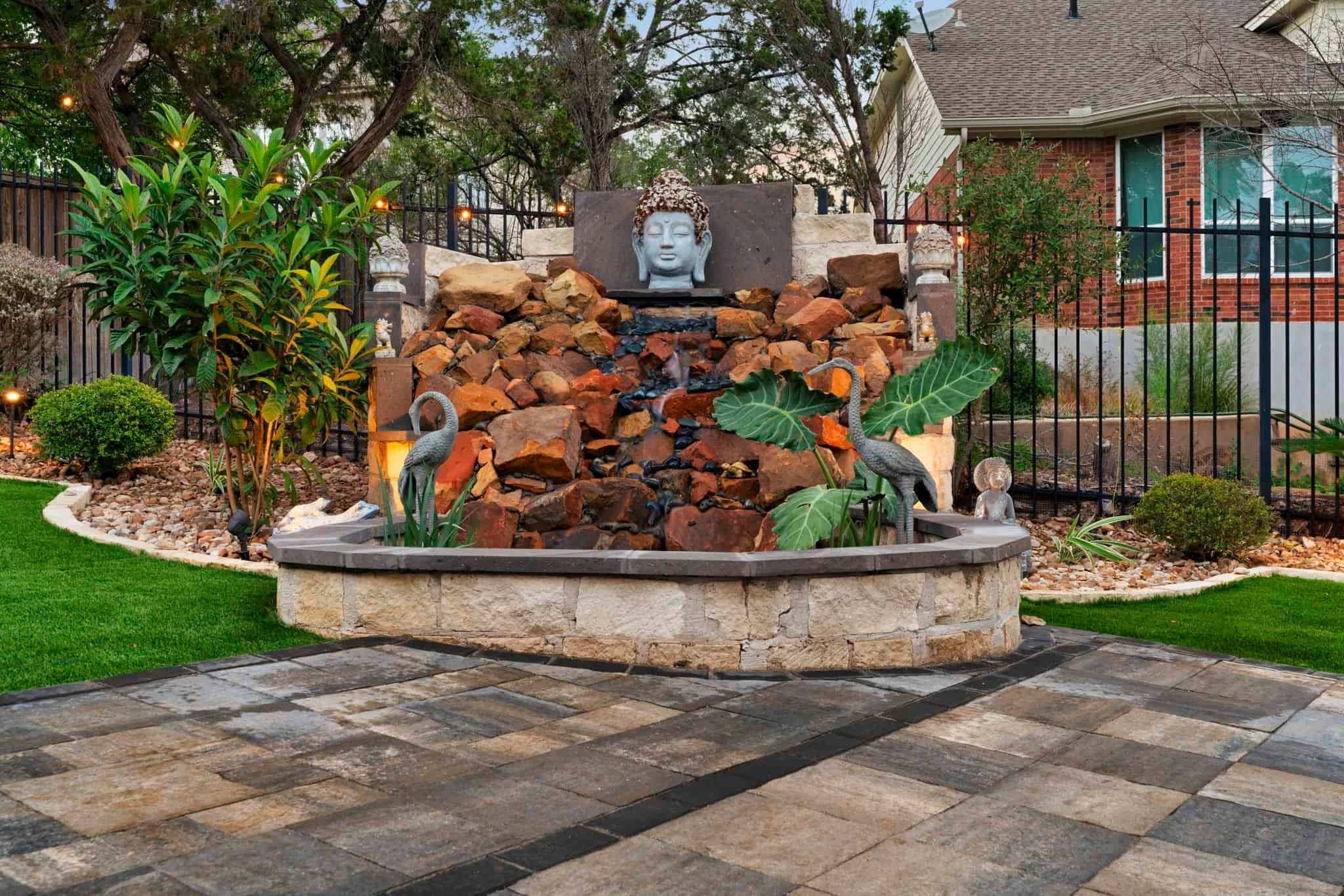 Decorative backyard water feature with stacked red rocks, a Buddha head sculpture, tropical plants, and crane statues.