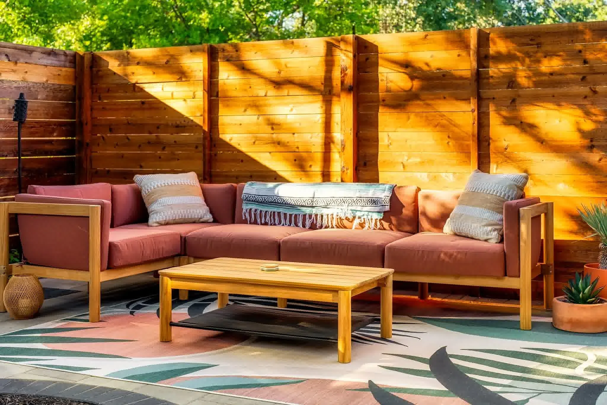 Modern wood privacy fence encloses a cozy patio with a pink cushioned sectional, an outdoor rug, and a coffee table, creating a stylish backyard retreat.