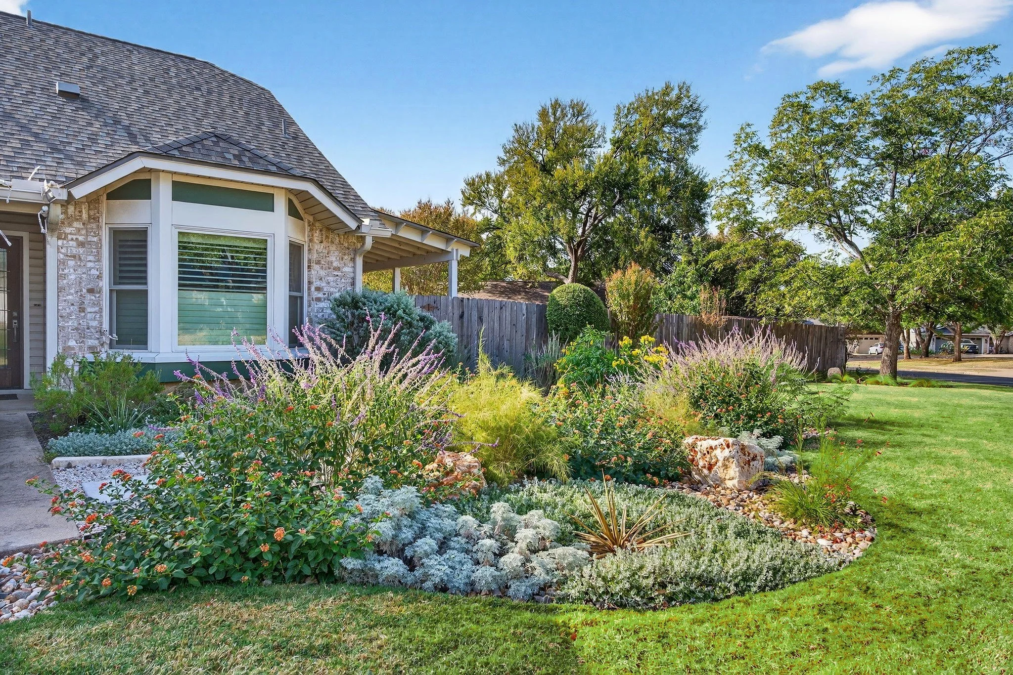 Curved perennial bed with lantana, artemisia, salvia, and limestone accent stones along a bay window