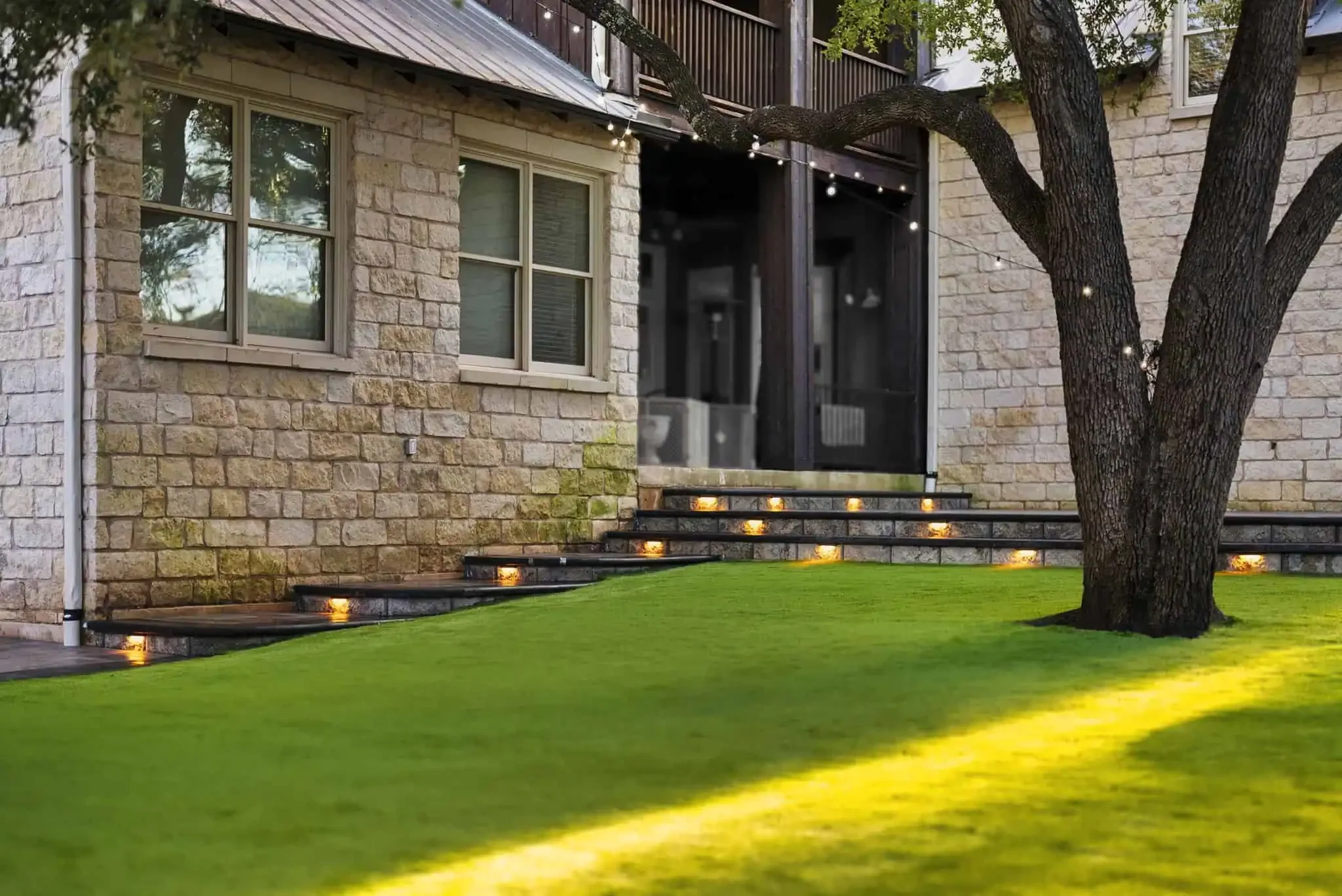 Modern outdoor living space with stone steps, soft lighting, green lawn, and a large tree outside a light stone house.