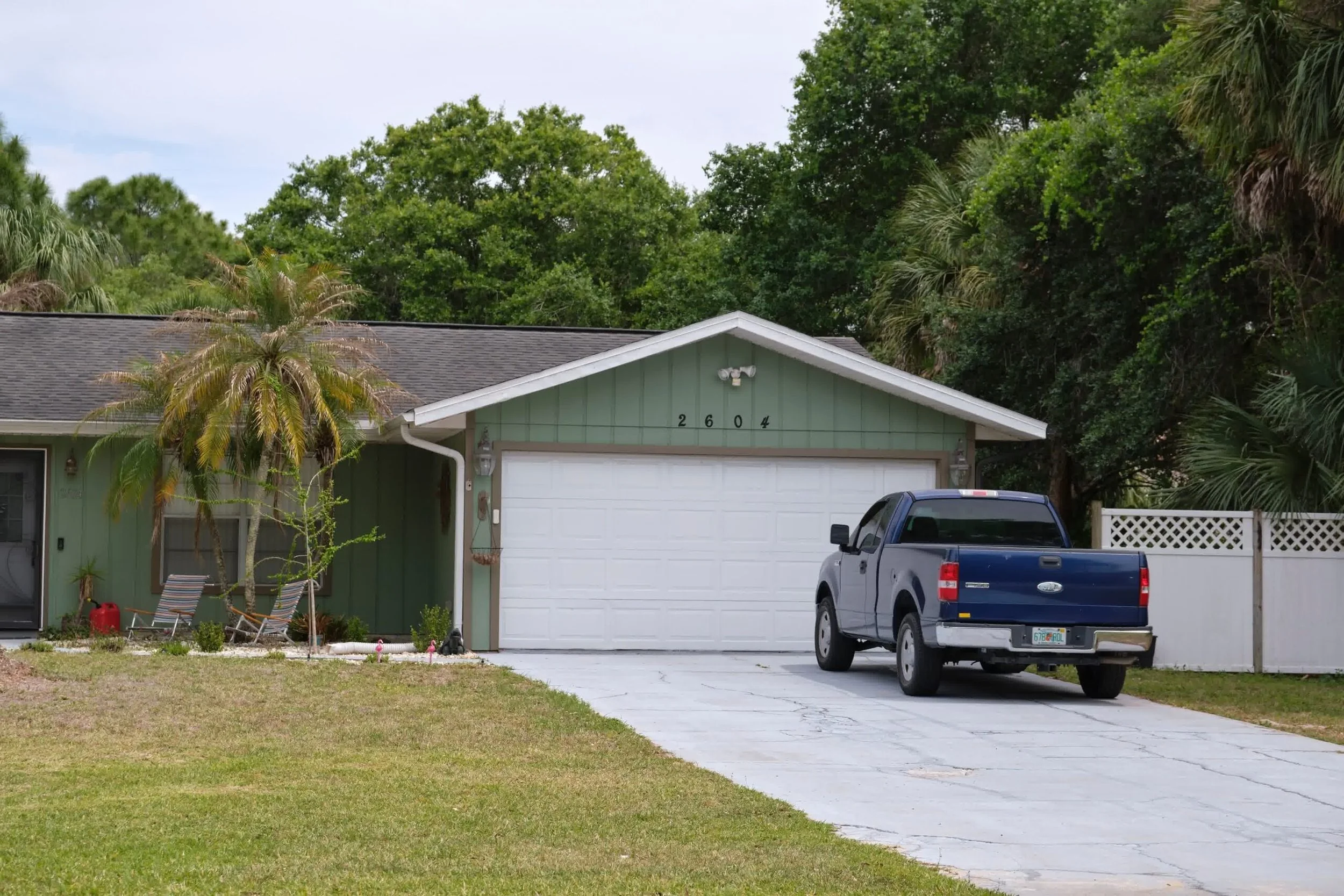 Single-family home with green siding, palm trees, and a concrete driveway leading to a two-car white garage in a suburban yard.
