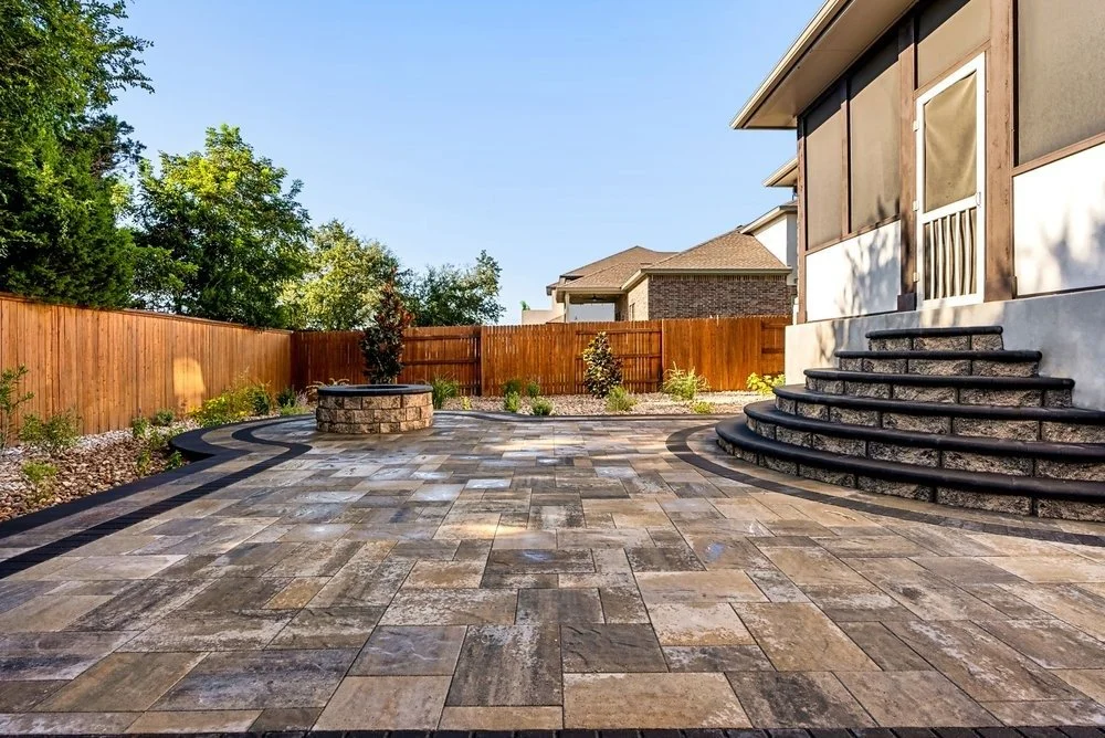 Spacious patio with multicolor stone pavers, curved steps, stone fire pit, and surrounding garden beds with wooden privacy fencing.