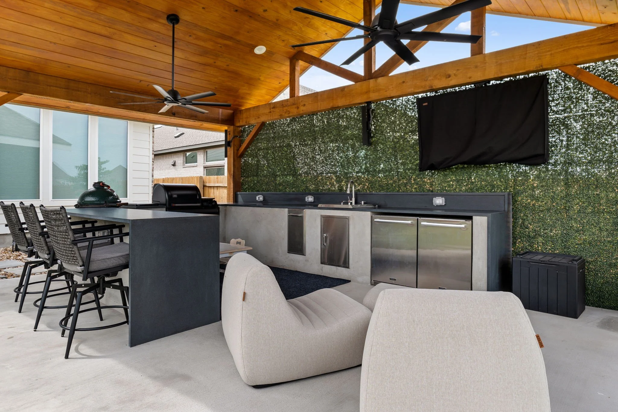 Outdoor kitchen with stainless grill, dual refrigerators, bar stools, and faux greenery privacy wall