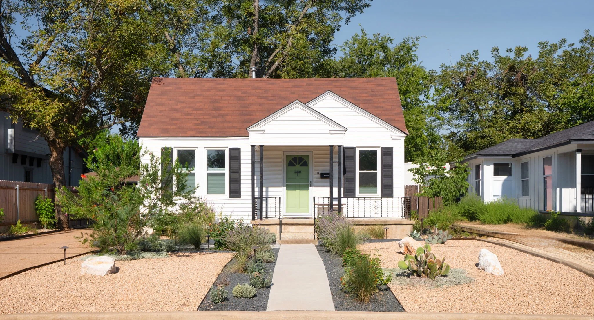 White cottage with green front door, concrete walkway, and xeriscaped yard of decomposed granite and native plants