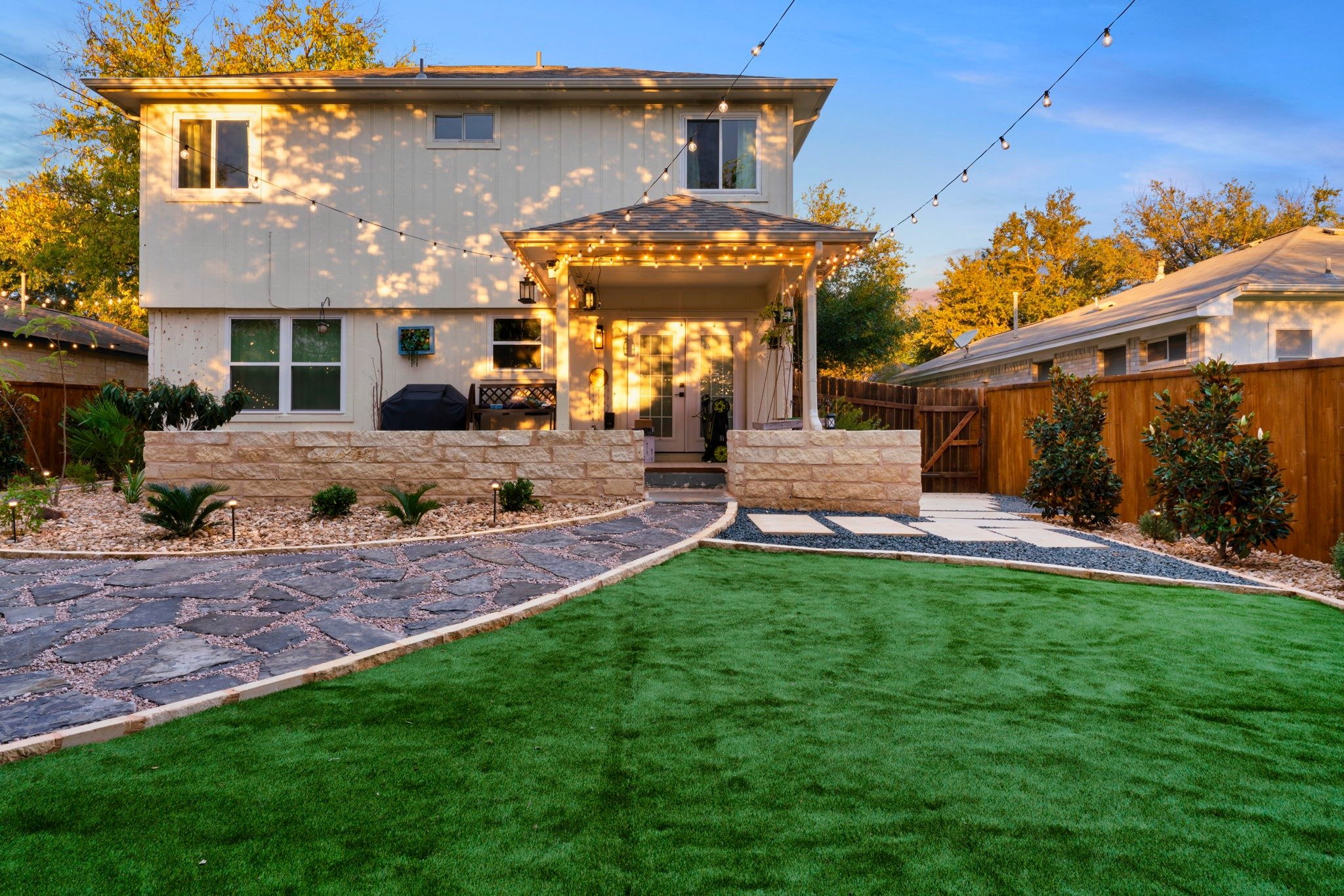 Backyard with artificial grass lawn, modern pergola, stone pathways, and warm lighting on a two-story cream house.