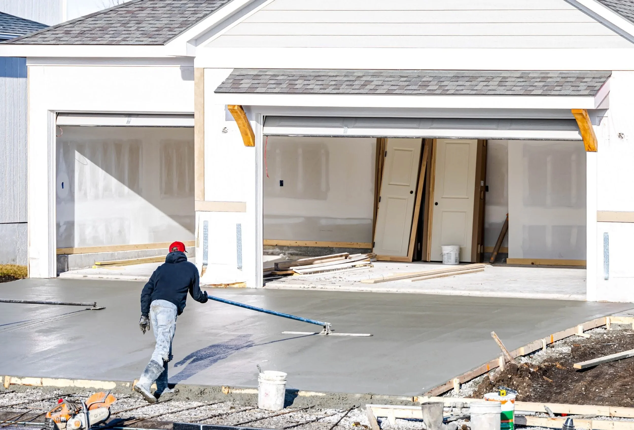 Contractor smooths freshly poured concrete driveway in front of new garage with a bull float at residential construction site.