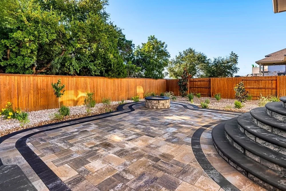 Backyard with curved stone paver patio, central fire pit, fresh landscaping, and a cedar privacy fence backed by lush green trees.
