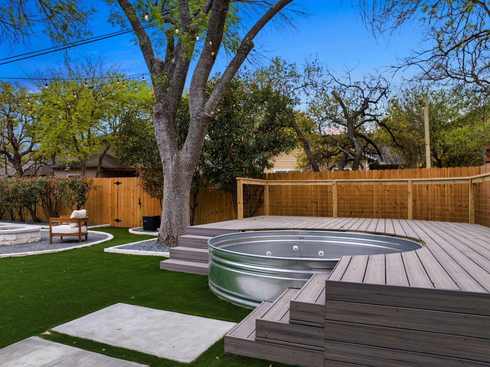 Backyard with a stock tank hot tub, wooden deck, artificial grass, and mature trees, enclosed by a wooden privacy fence.
