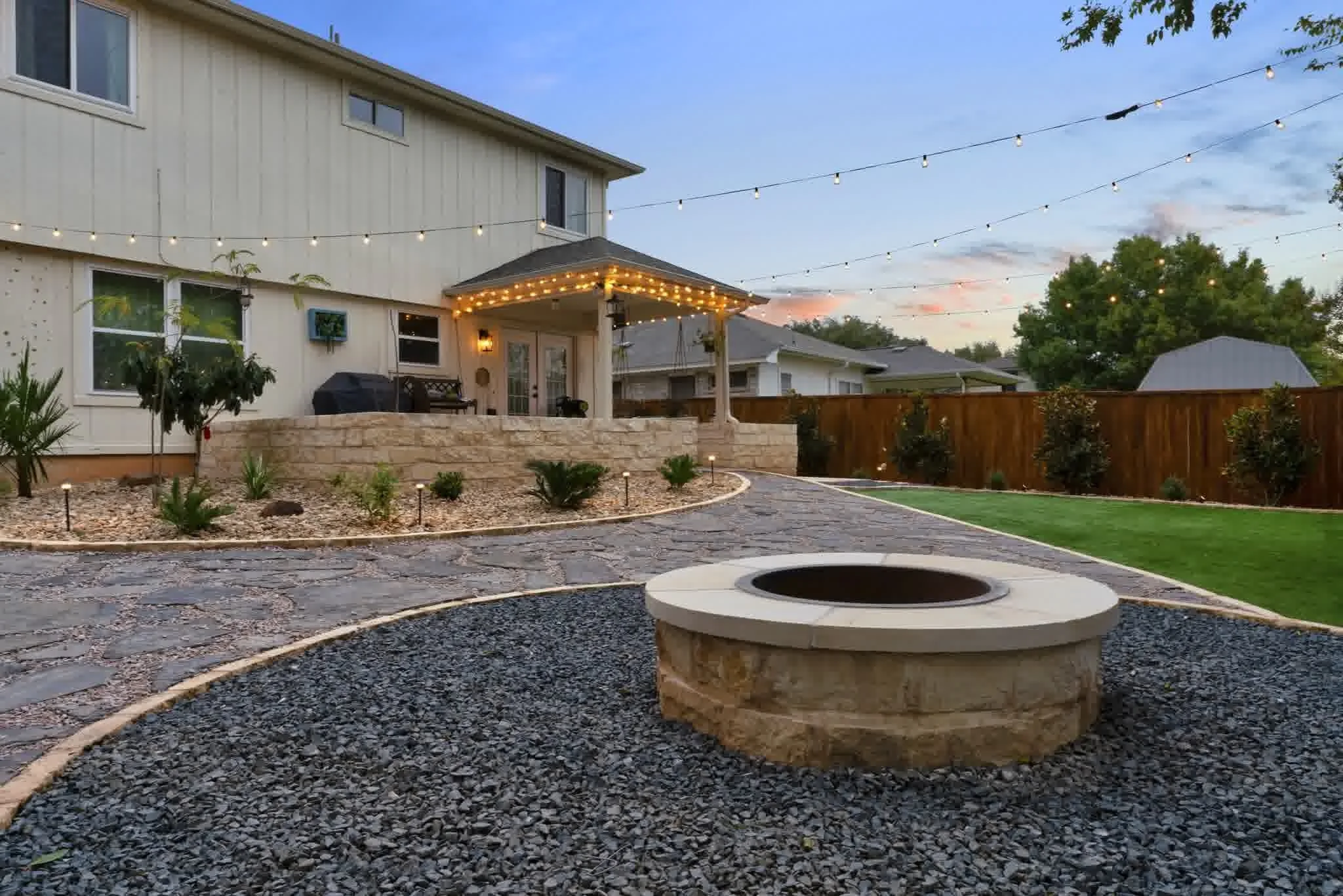 Stone firepit on gravel patio with string lights, landscaped garden beds, and covered porch seating in landscaped backyard at sunset.