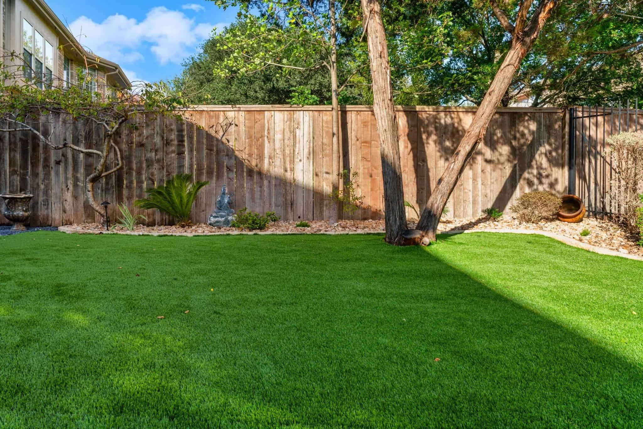 Backyard with vibrant artificial turf, wooden privacy fence, trees, decorative plants, and a Buddha statue near a stone border.