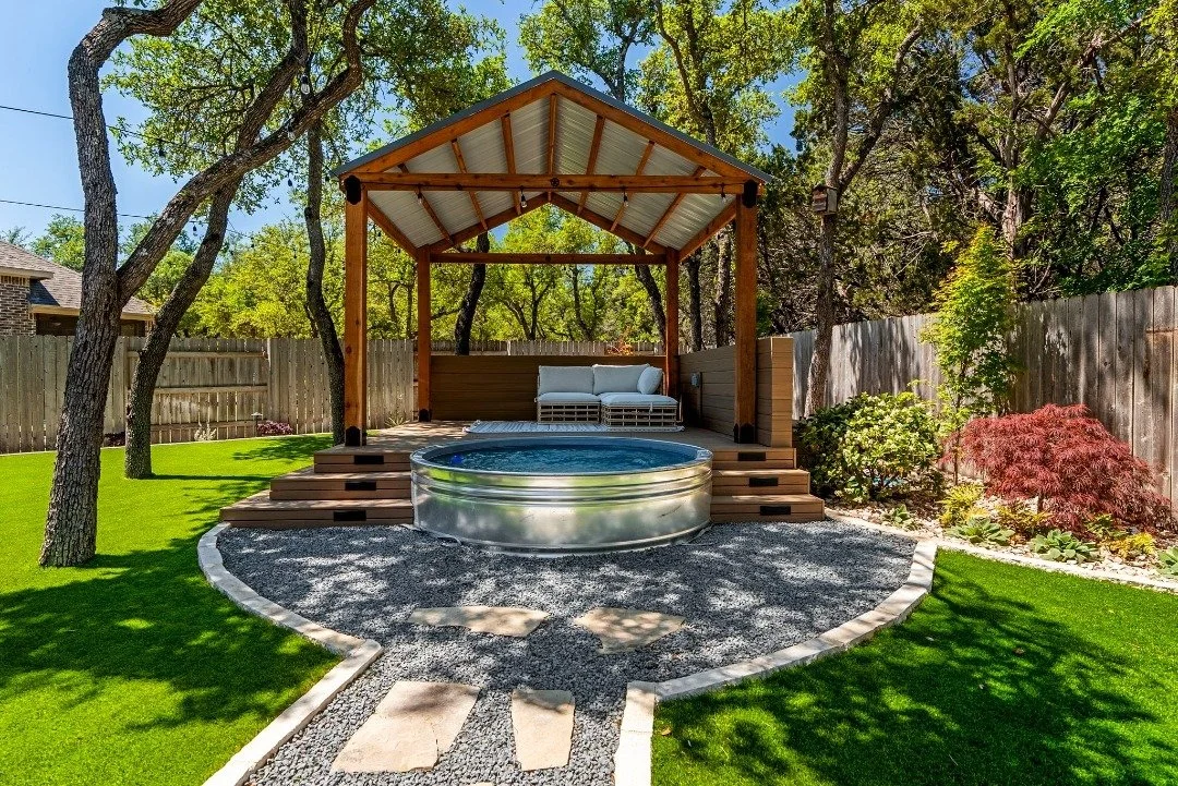 Backyard oasis with a stock tank pool, wooden pergola, outdoor lounge seating, and lush artificial grass surrounded by trees.